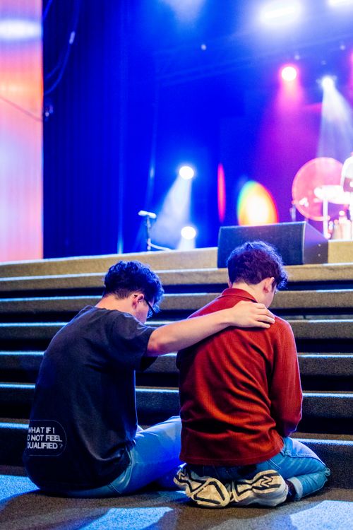Free Two young men in a supportive embrace during an indoor event, enhanced by colorful stage lighting. Stock Photo