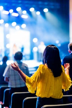 A woman in a yellow blouse with hands raised at a lively concert event indoors.