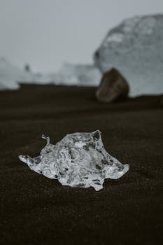 A crystalline ice shard rests on dark, volcanic sand, creating a stark contrast in color and texture.