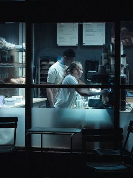 Two baristas working in a cafe at night, seen through a window with a dimly lit ambiance.