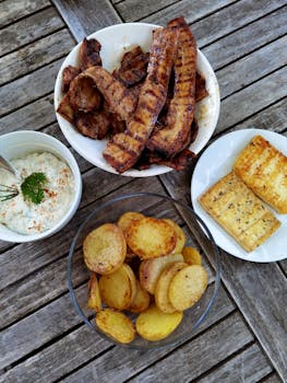 Delicious outdoor barbecue featuring grilled pork, potatoes, tofu, and a creamy dip on a rustic table.