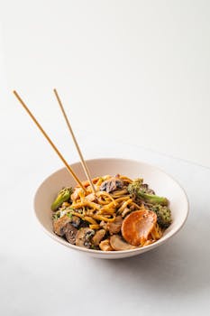 A delectable bowl of Asian-style stir fry noodles with mushrooms, broccoli, and chopsticks.