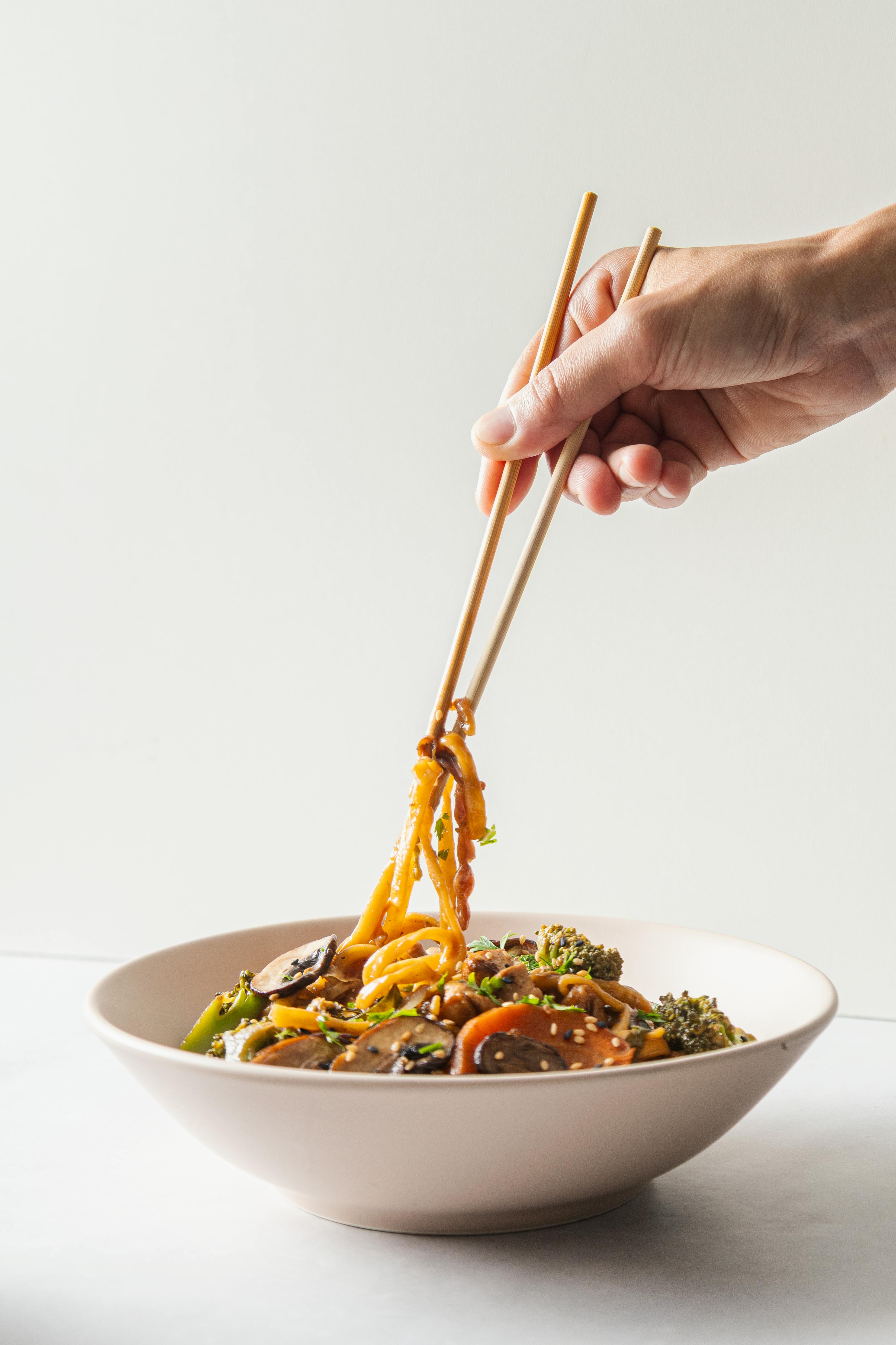 Vibrant stir fry with vegetables and noodles being picked up with chopsticks against a light background.