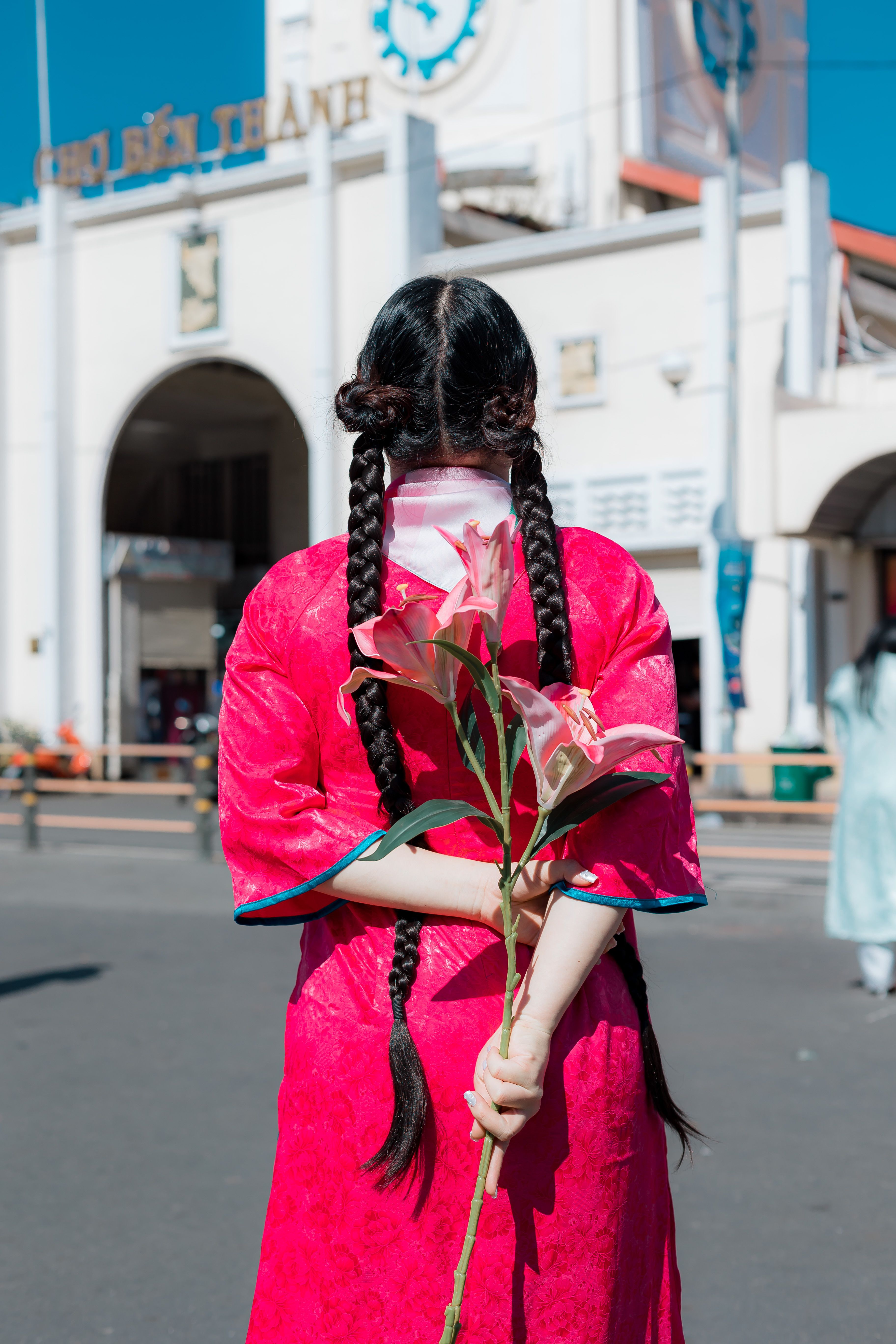 Back view of a woman in traditional dress holding a pink lily at a market entrance.
