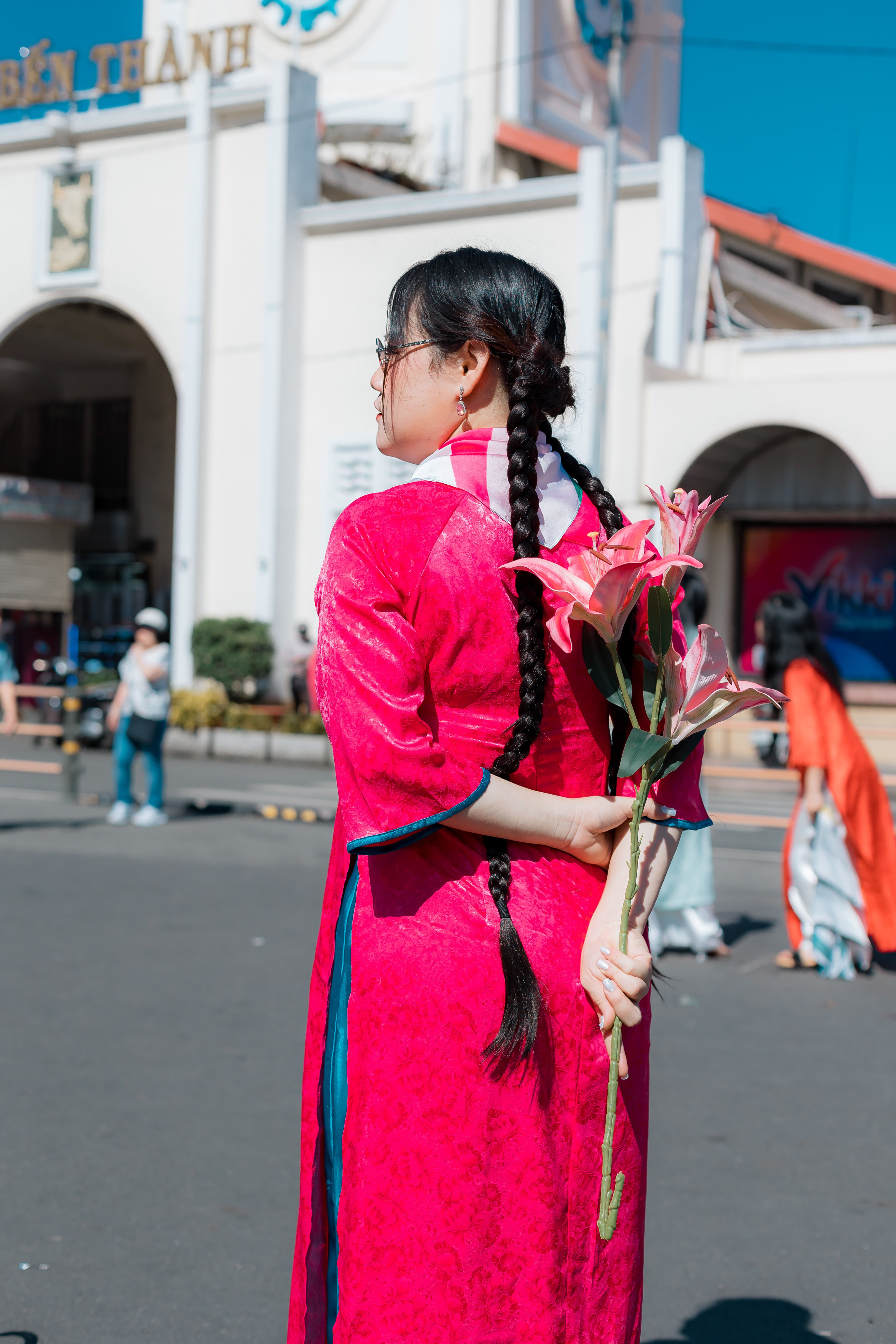 Woman in pink traditional dress holds lilies at Ben Thanh Market outdoor setting.