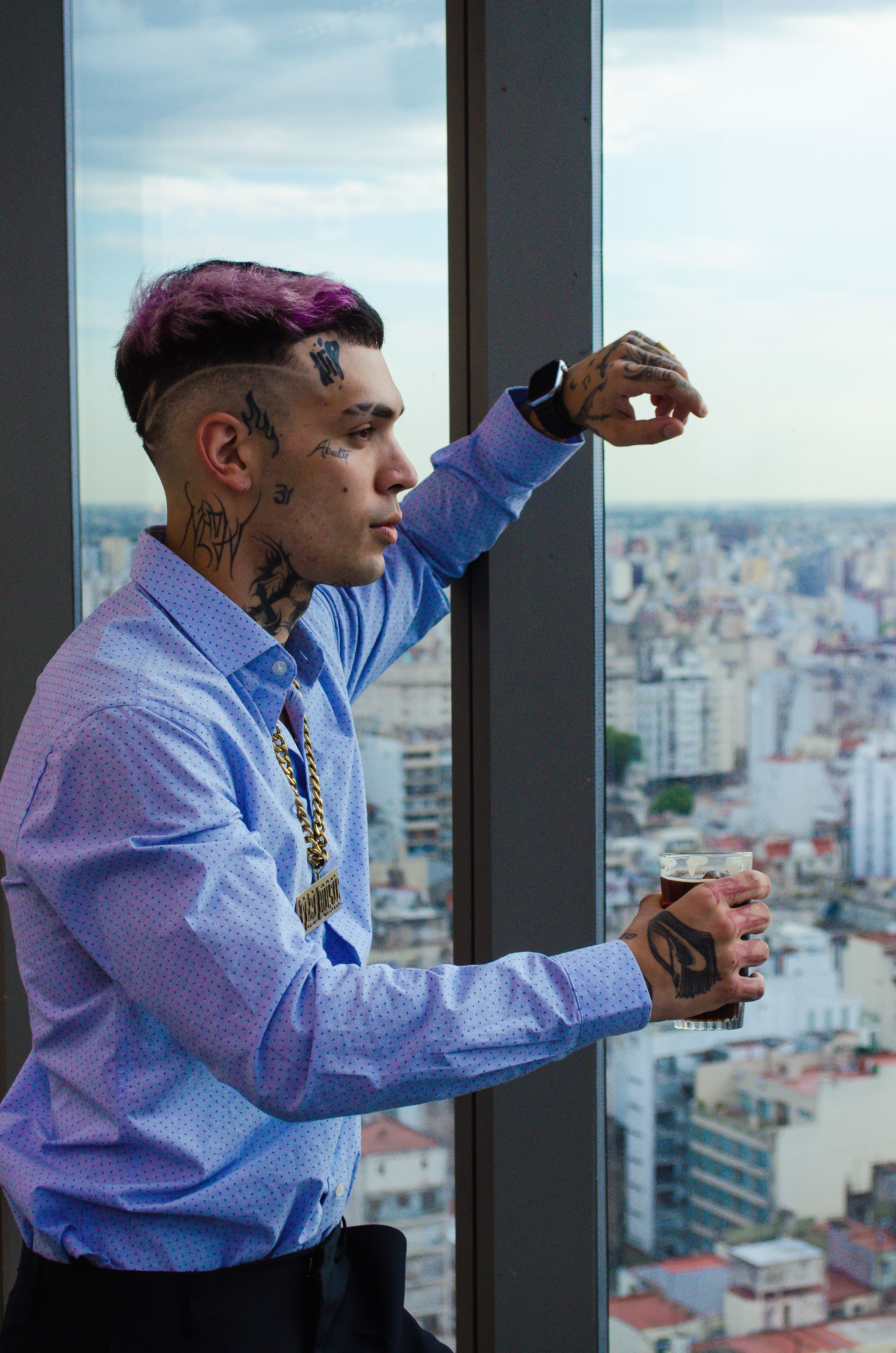 Young man with tattoos and dyed hair gazes out from high-rise window overlooking cityscape.