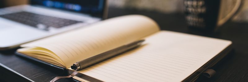 Open notebook with a pen on a wooden desk beside a laptop, ideal for blogging and work-related themes.