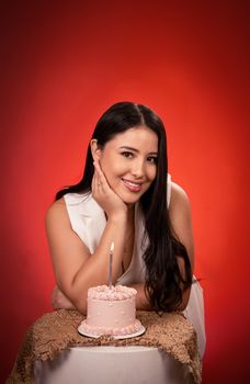 Smiling woman in white beside a small pink birthday cake with a candle.
