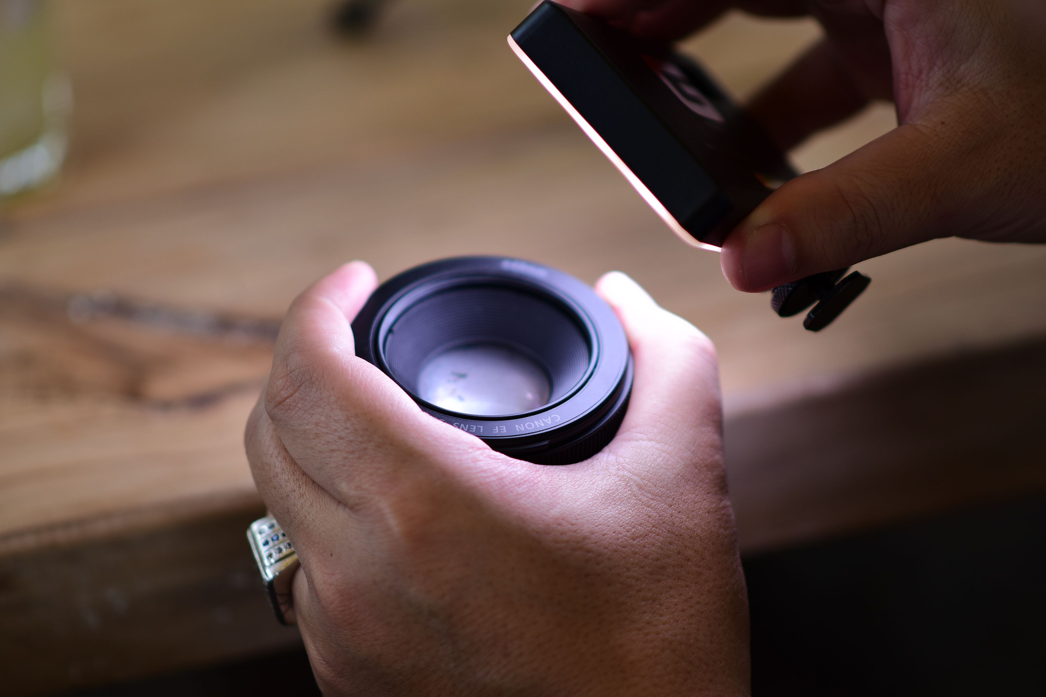 A detailed view of hands examining a camera lens under a light source on a wooden table.