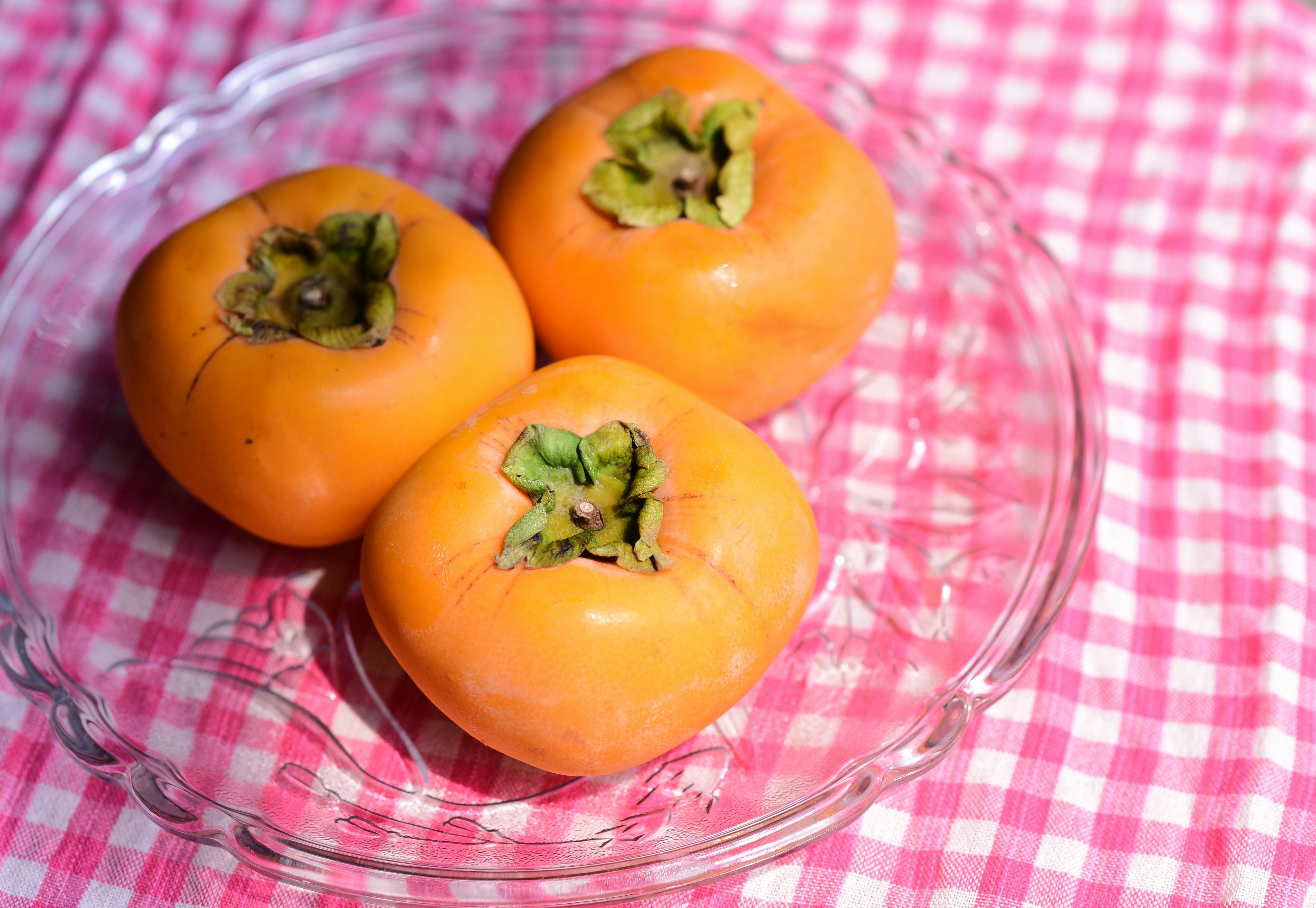 Three ripe persimmons beautifully displayed in a glass dish on a pink checkered tablecloth.