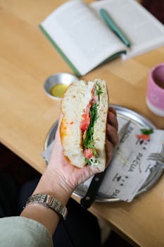 A fresh veggie sandwich being held, with book and coffee in a cozy indoor setting.