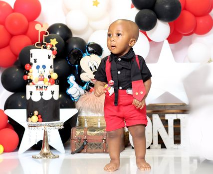 A cute baby enjoying a Mickey Mouse themed birthday party with colorful balloons and cake.