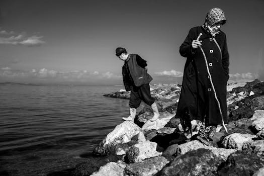 Black and white photo of elderly couple navigating rocky shoreline by the sea.