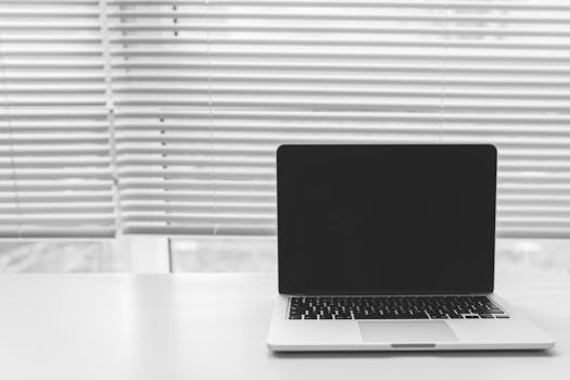 Black and white photo of a laptop on a white desk with window blinds in the background.