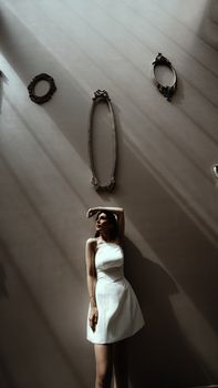 A stylish woman in a white dress poses dramatically in shadow against a wall with vintage frames.