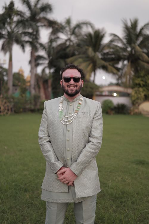 Free A man in a stylish traditional outfit stands outdoors with palm trees in the background. Stock Photo