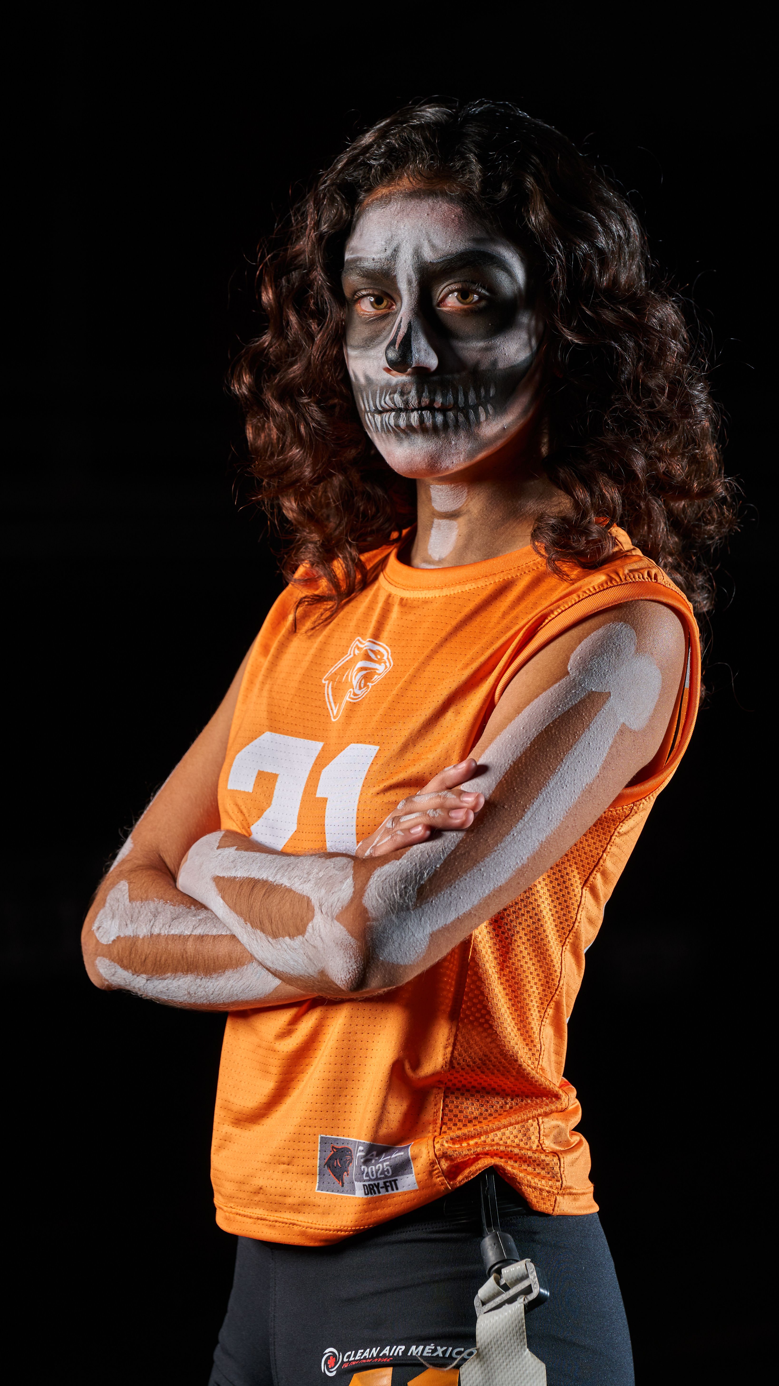 Portrait of a female athlete in skeleton makeup wearing an orange jersey, posing confidently with arms crossed.