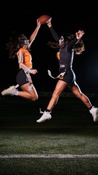 Two women in face paint leap for football at night on field.
