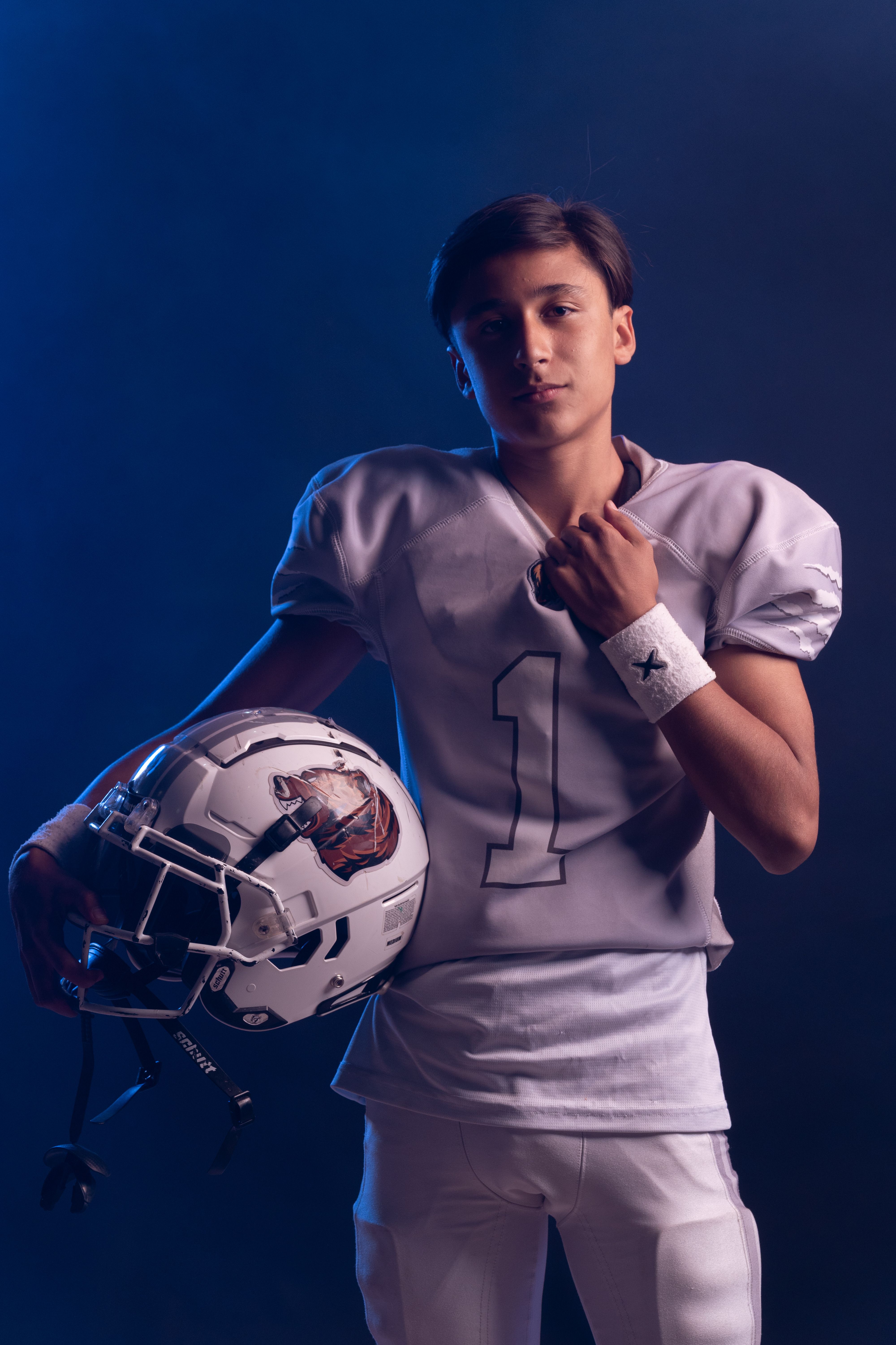 Teen football player in uniform poses with helmet on dark background.