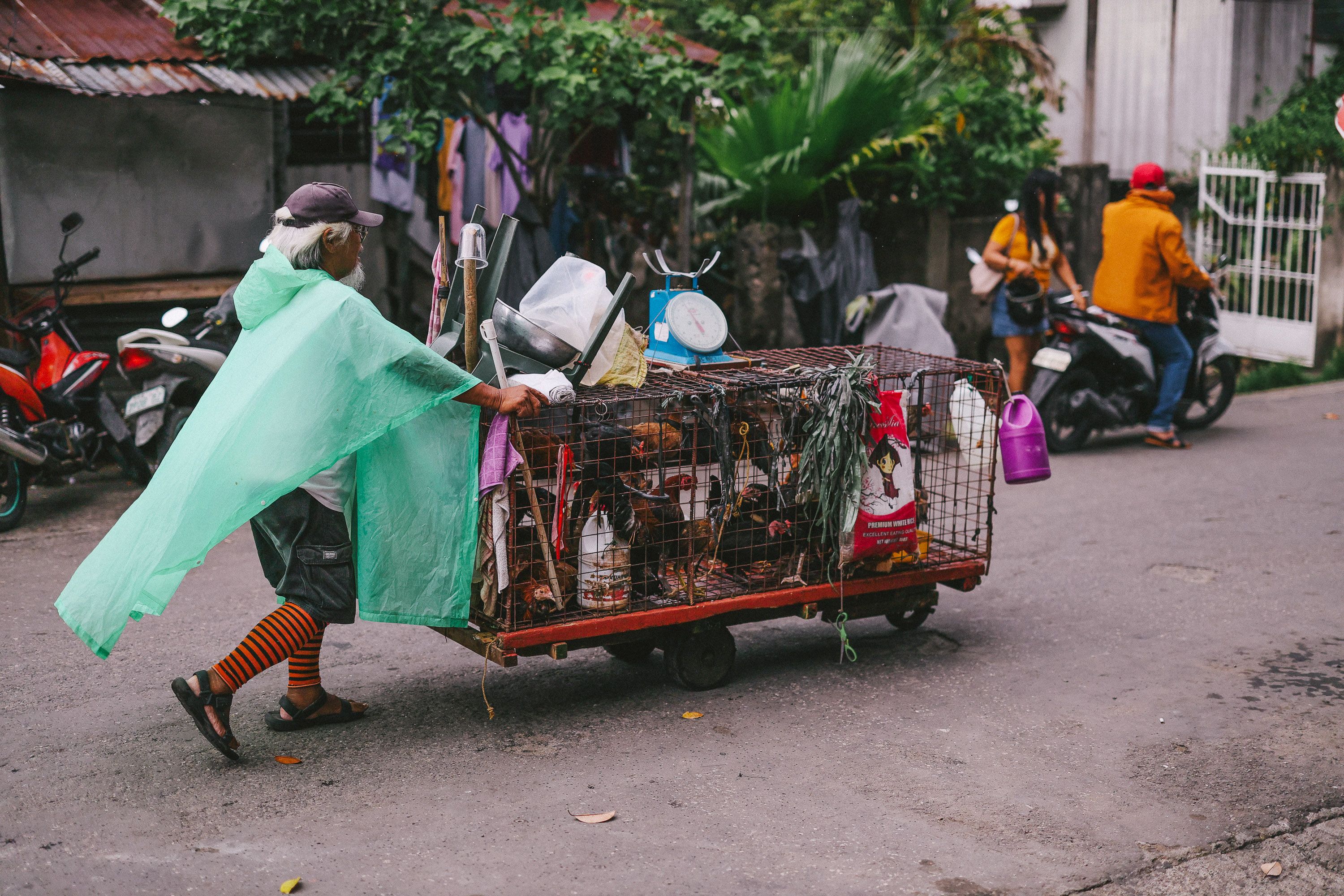 Older man pushing a cart full of recyclables on a city street wearing a green rain poncho.
