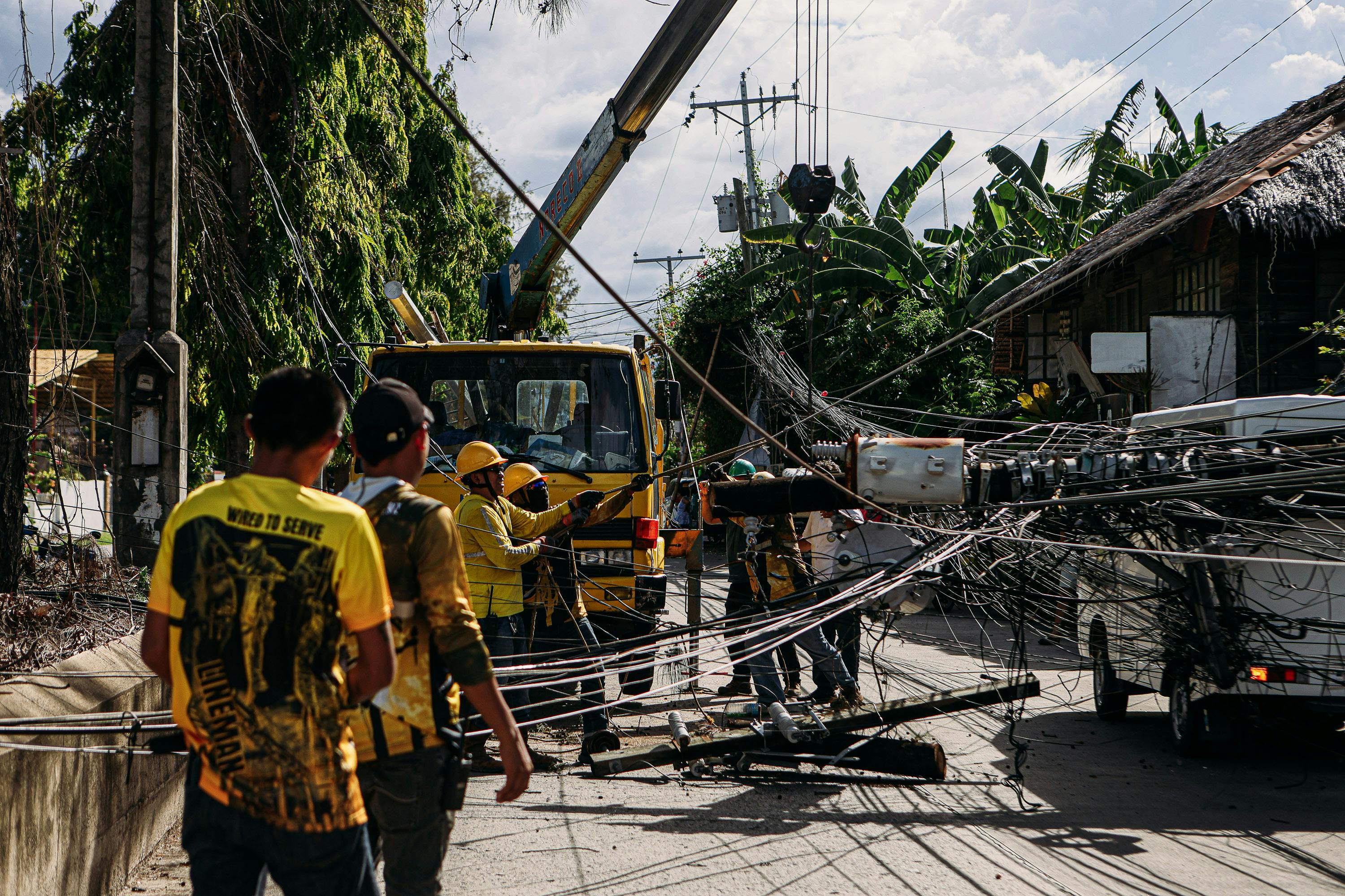 Workers fixing damaged electrical lines on a street post-storm, surrounded by fallen cables and equipment.