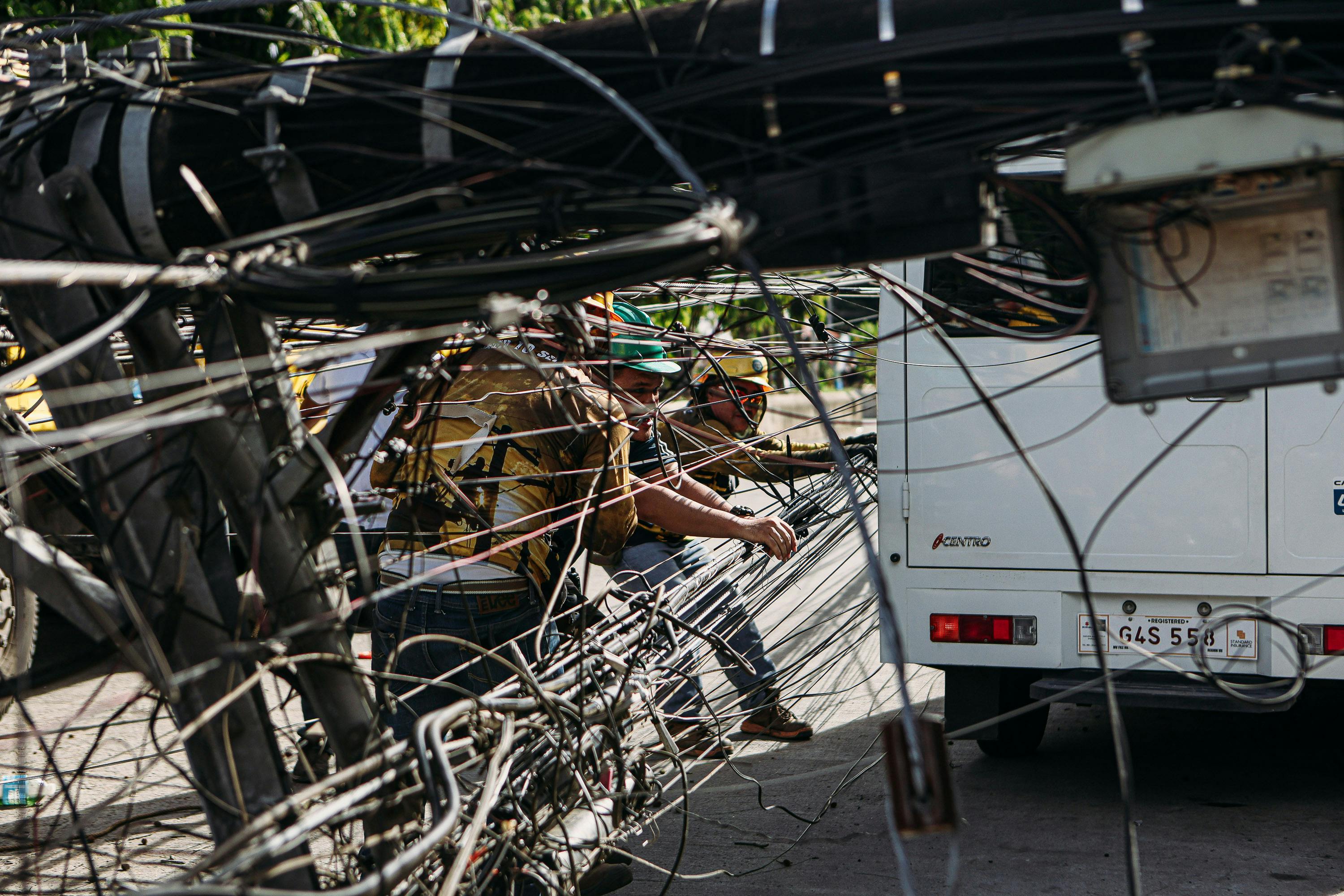 Workers managing tangled power lines by a street setup near a vehicle, highlighting infrastructure challenges.
