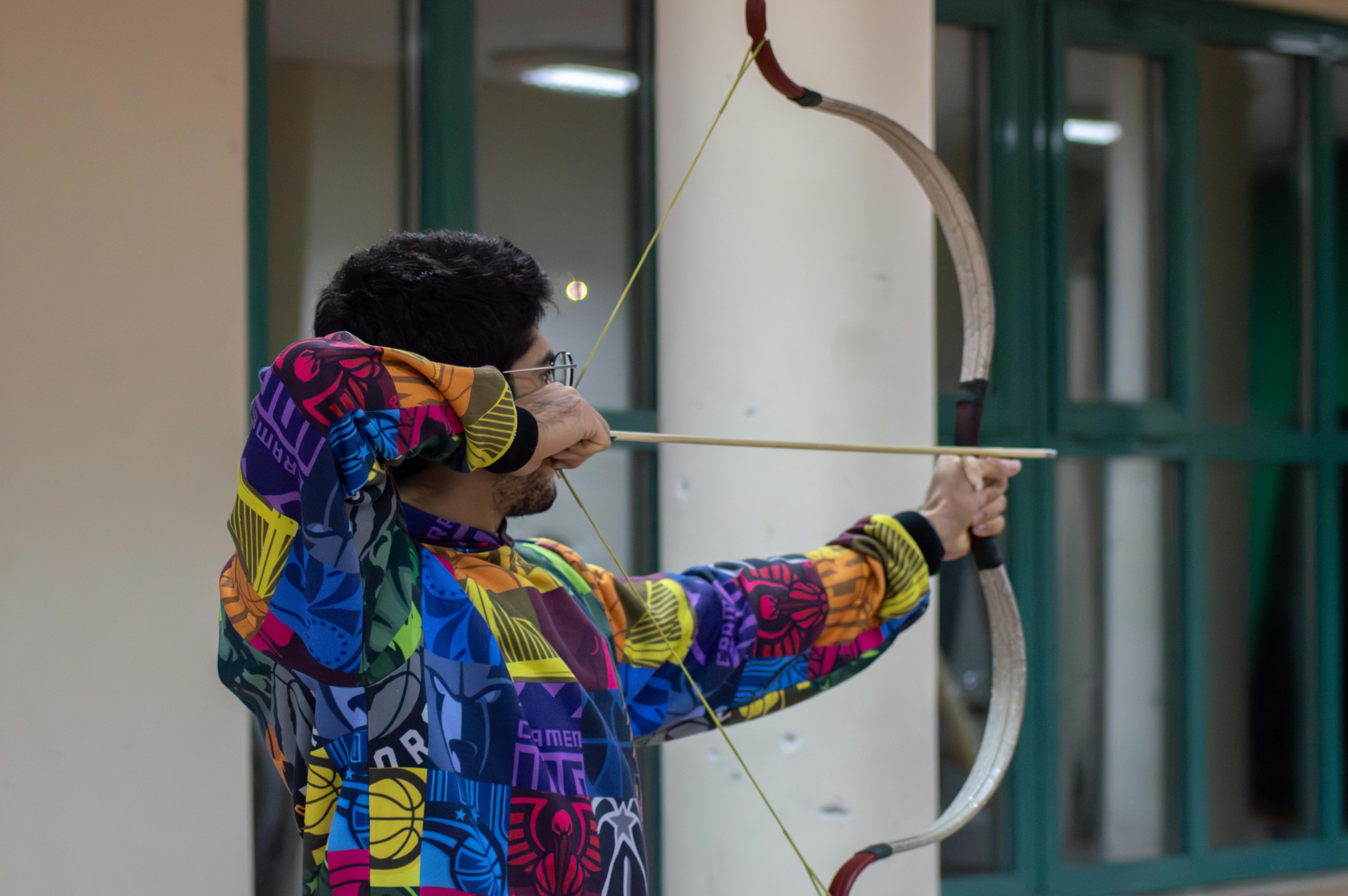A man in a colorful jacket practicing archery indoors with a focus on aiming.