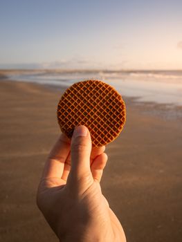 Hand holding a stroopwafel in front of IJmuiden beach during sunset, capturing a peaceful outdoor moment.