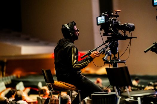 A focused cameraman operates a video camera on a tripod in a dimly lit indoor setting.