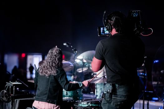 A drummer performs on stage while a cameraman captures the live event.