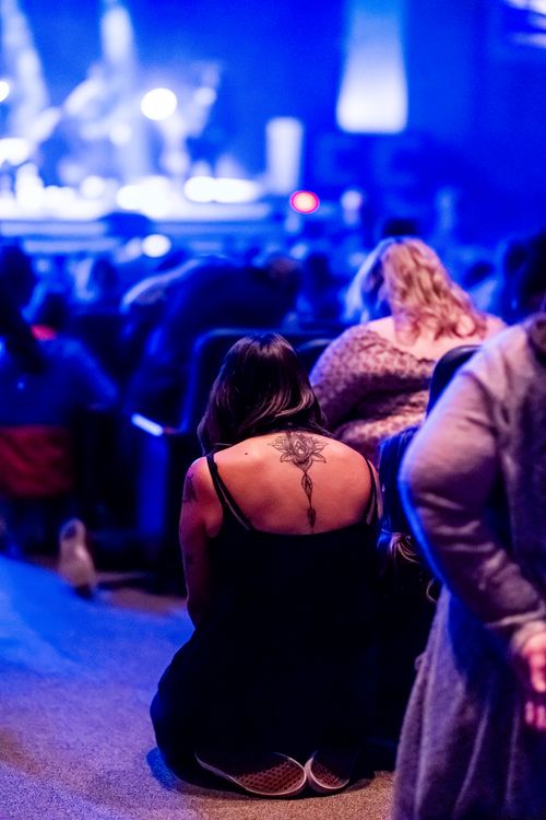 Free A woman with a tattoo kneels in prayer amidst a vibrant concert audience. Stock Photo