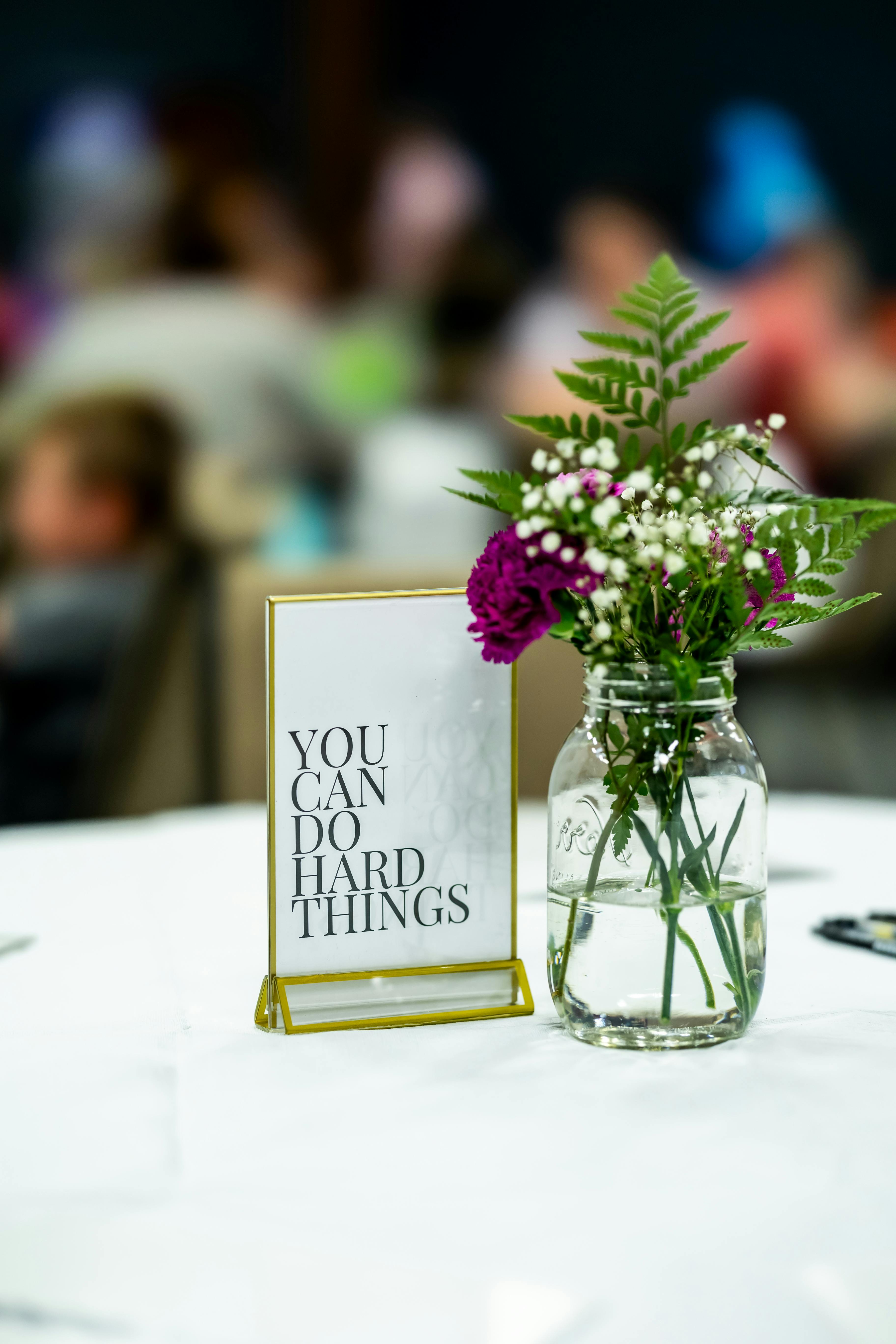 A motivational sign beside a floral arrangement in a mason jar at a gathering.