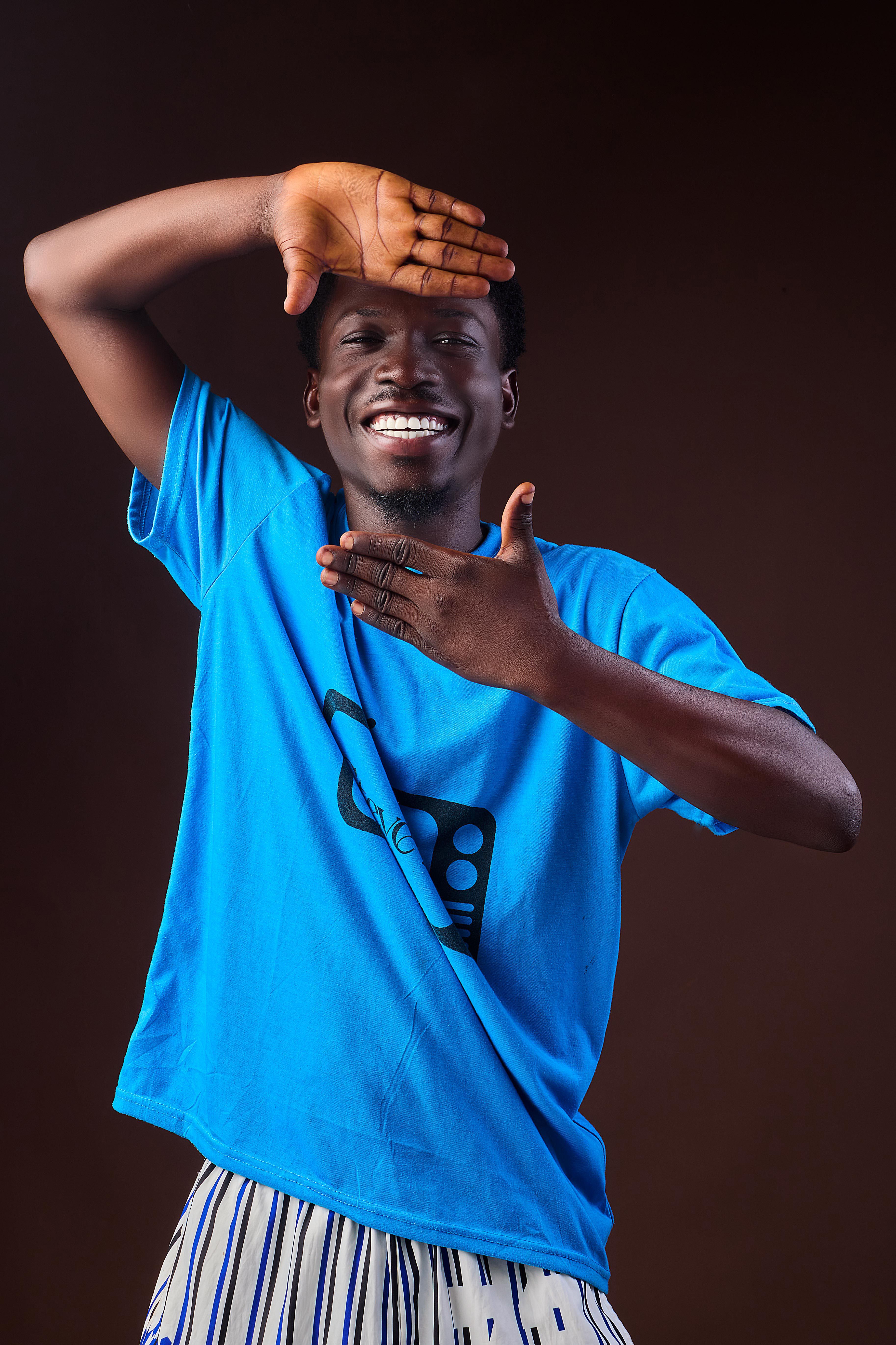 Cheerful young man in blue shirt, smiling and posing playfully indoors with a dark background.