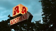 Berlin pharmacy sign with iconic logo during twilight, surrounded by trees.