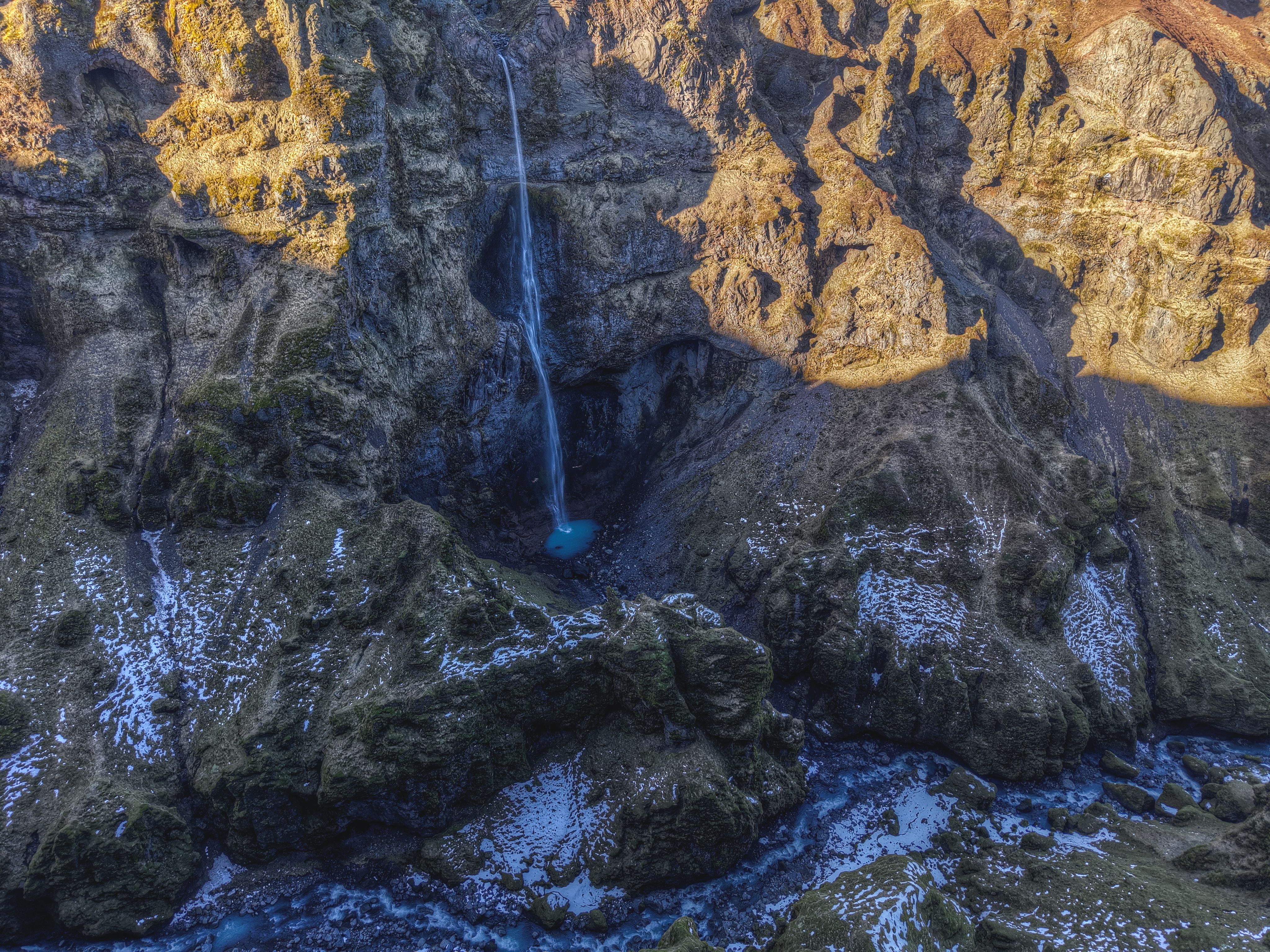 Best Helicopter Tour: Reykjavik City with landing at Mount Esja Review Review 2026 - A mesmerizing aerial view of a waterfall cascading into a pool, surrounded by snowy Icelandic cliffs.