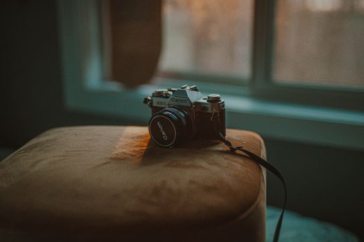A Canon vintage camera rests on a stool by a window with soft sunlight filtering through.