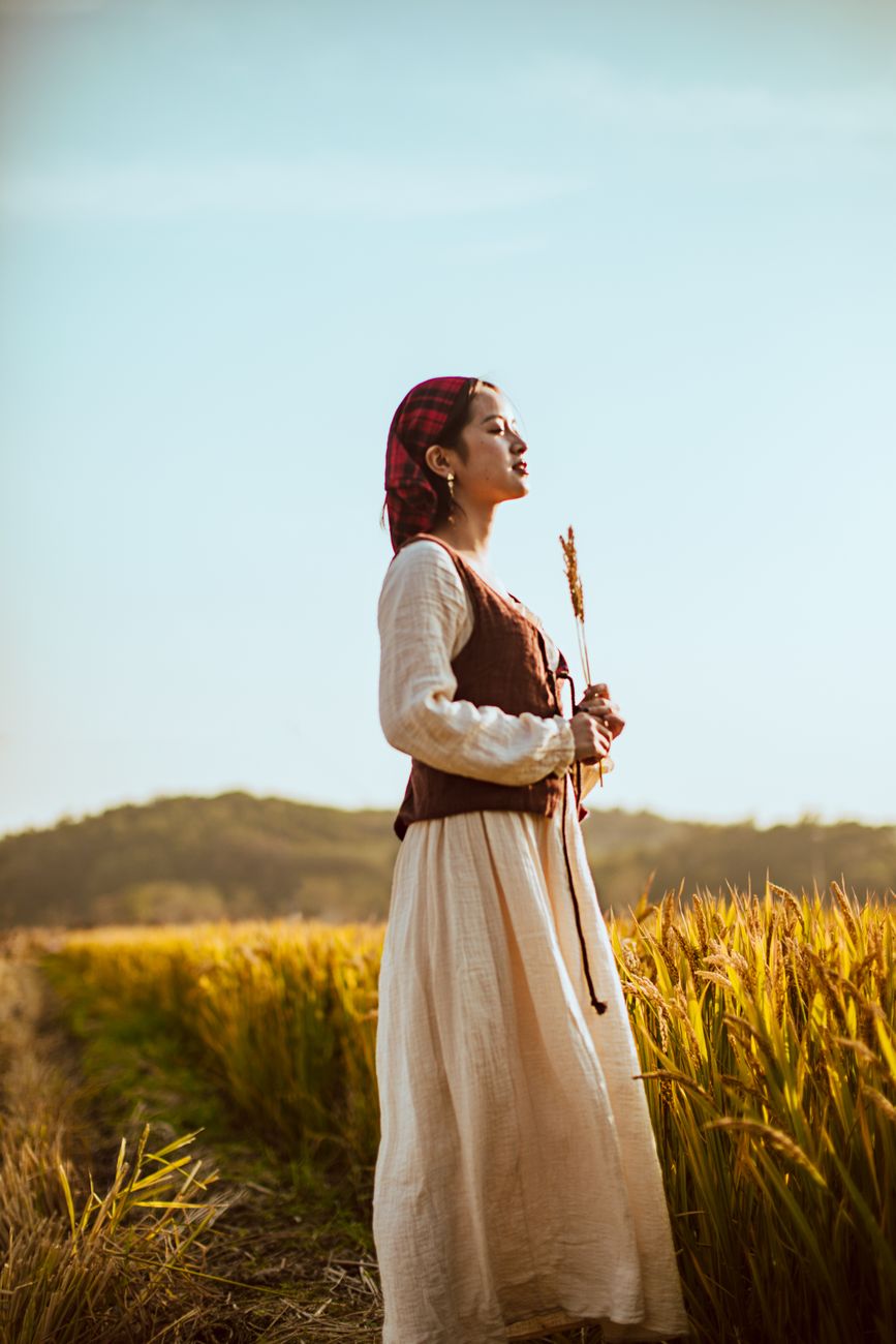 A woman in traditional attire stands in a wheat field under clear skies, embodying pastoral tranquil