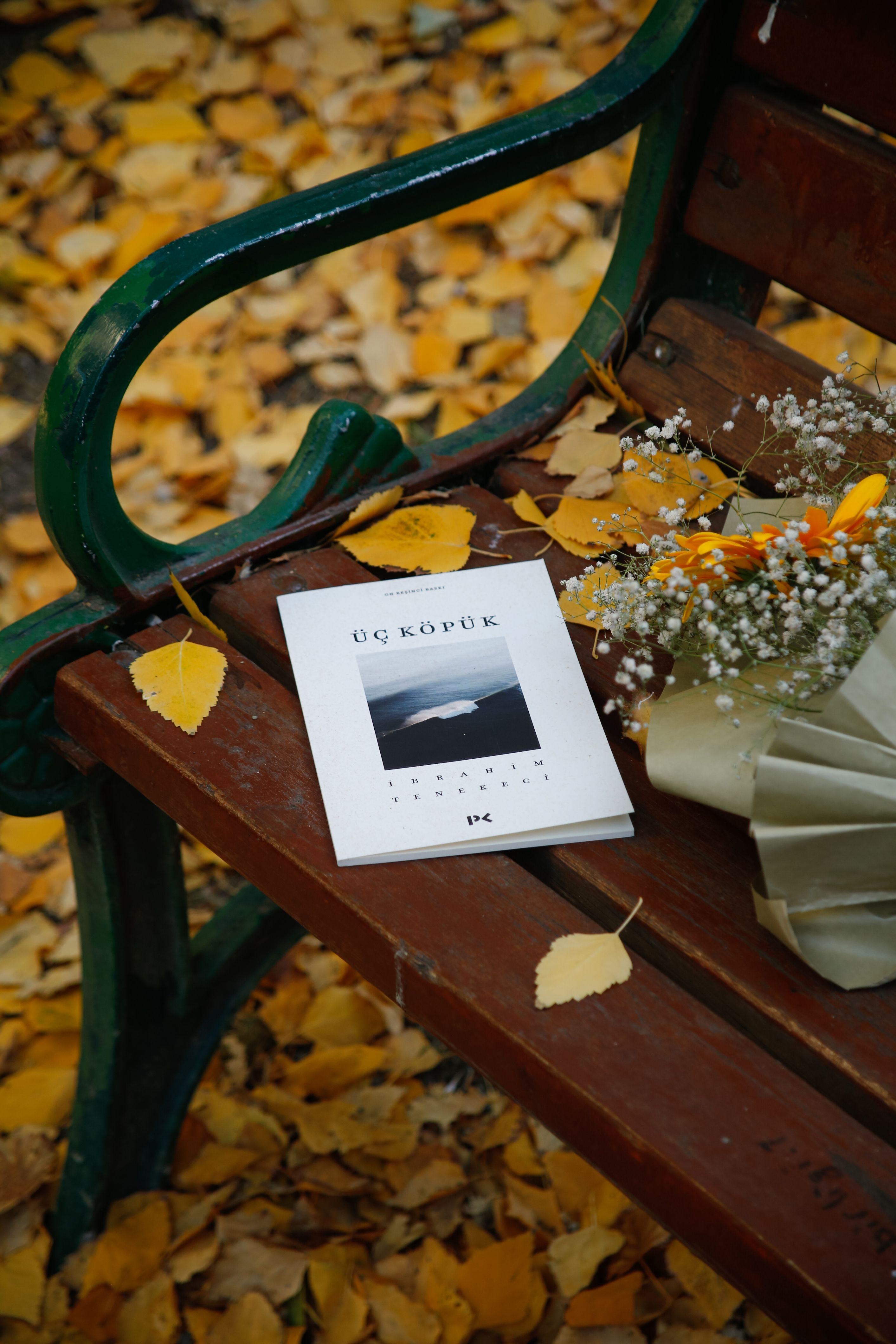 A serene autumn scene with a book and flowers on a park bench surrounded by fallen leaves.