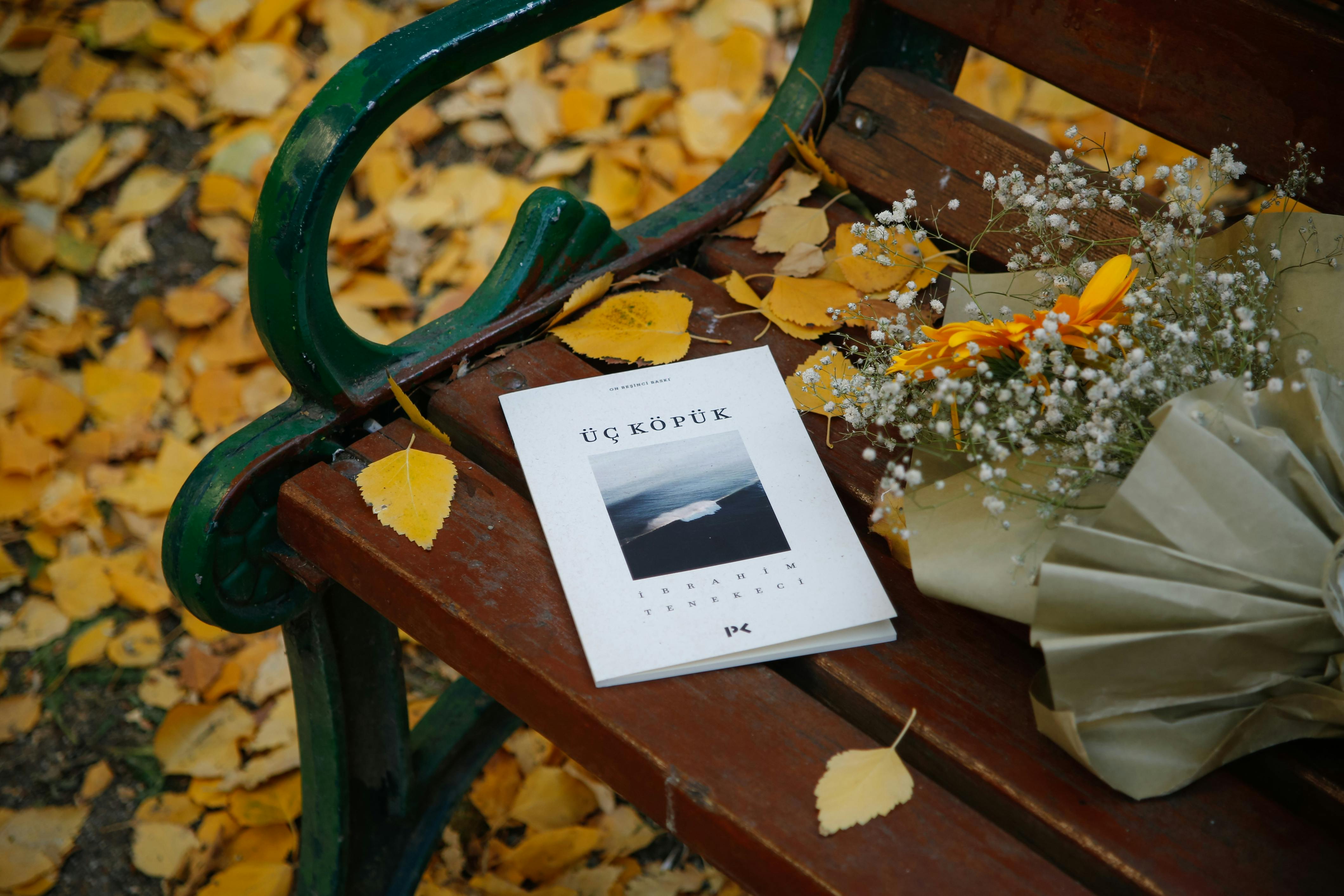 A book and flowers on a bench surrounded by autumn leaves in a park.