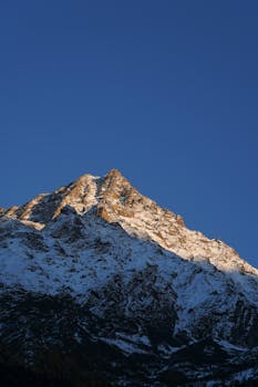 Snowy mountain peak glistens under a clear blue sky at sunset, capturing nature's beauty.
