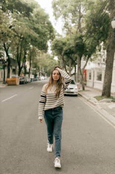 A woman walking confidently down a tree-lined urban street, showcasing street fashion.
