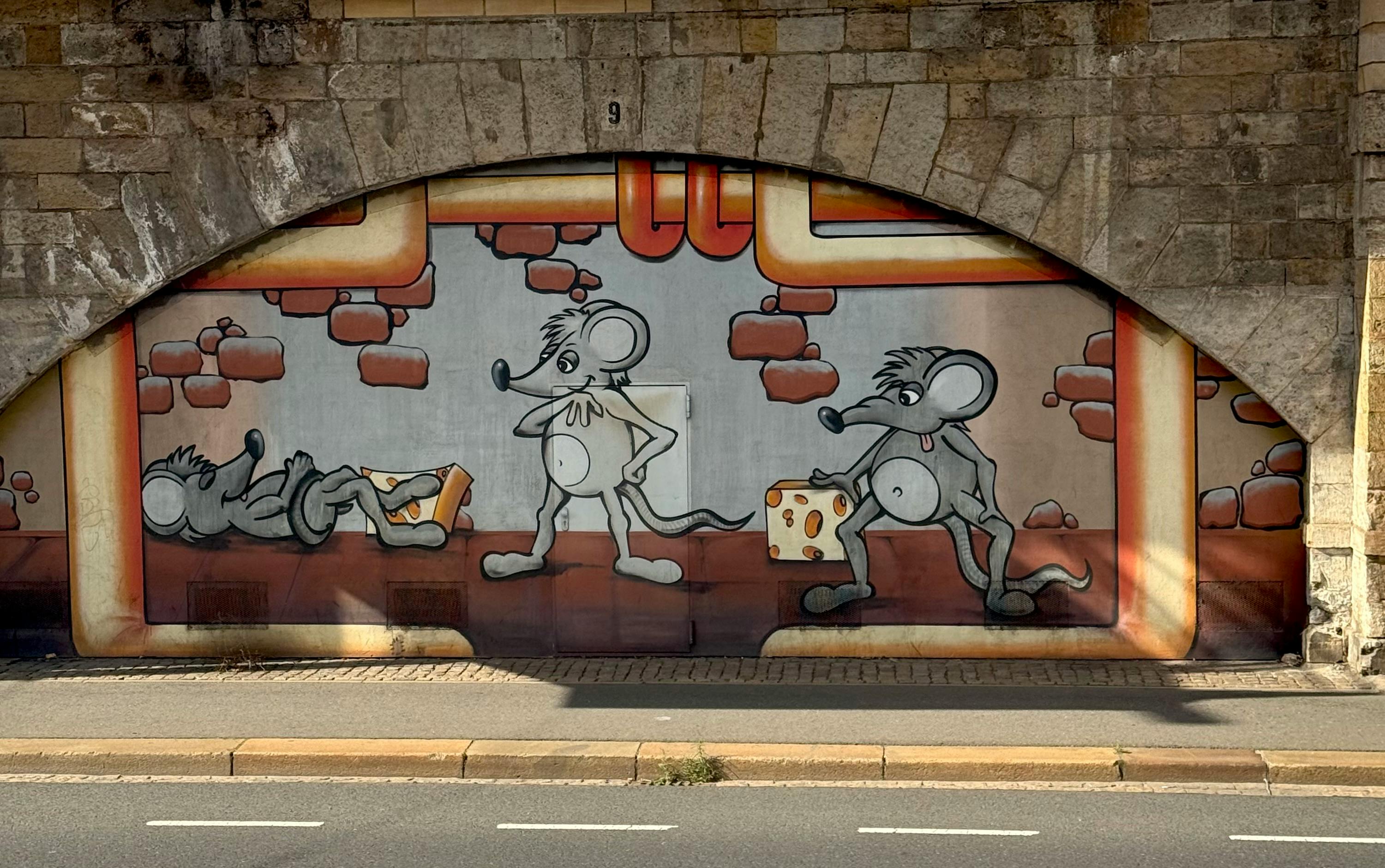 Colorful mural of cartoon mice with cheese on a wall under an arch in Germany.