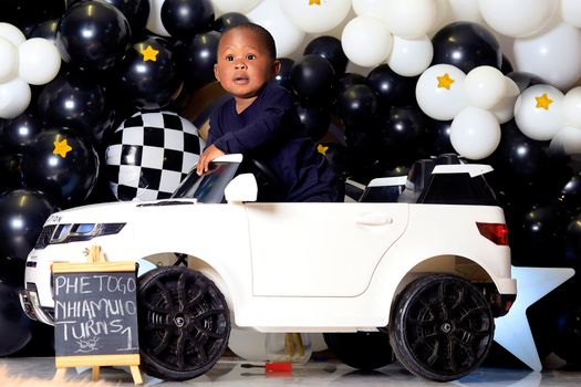Adorable baby boy having fun in a toy car at his first birthday party with a balloon backdrop.