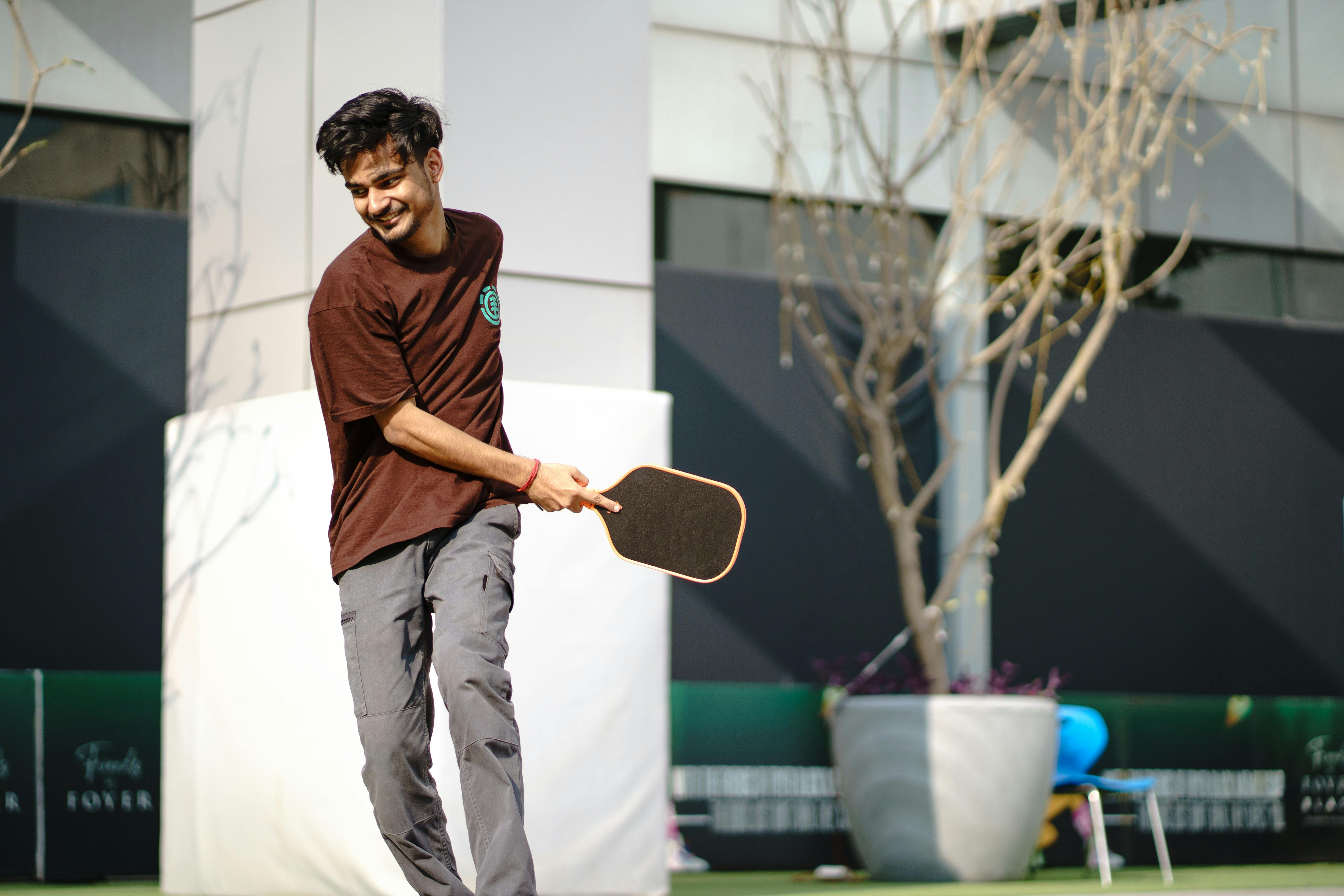 A smiling young man actively plays pickleball on an outdoor court, enjoying a sunny day.