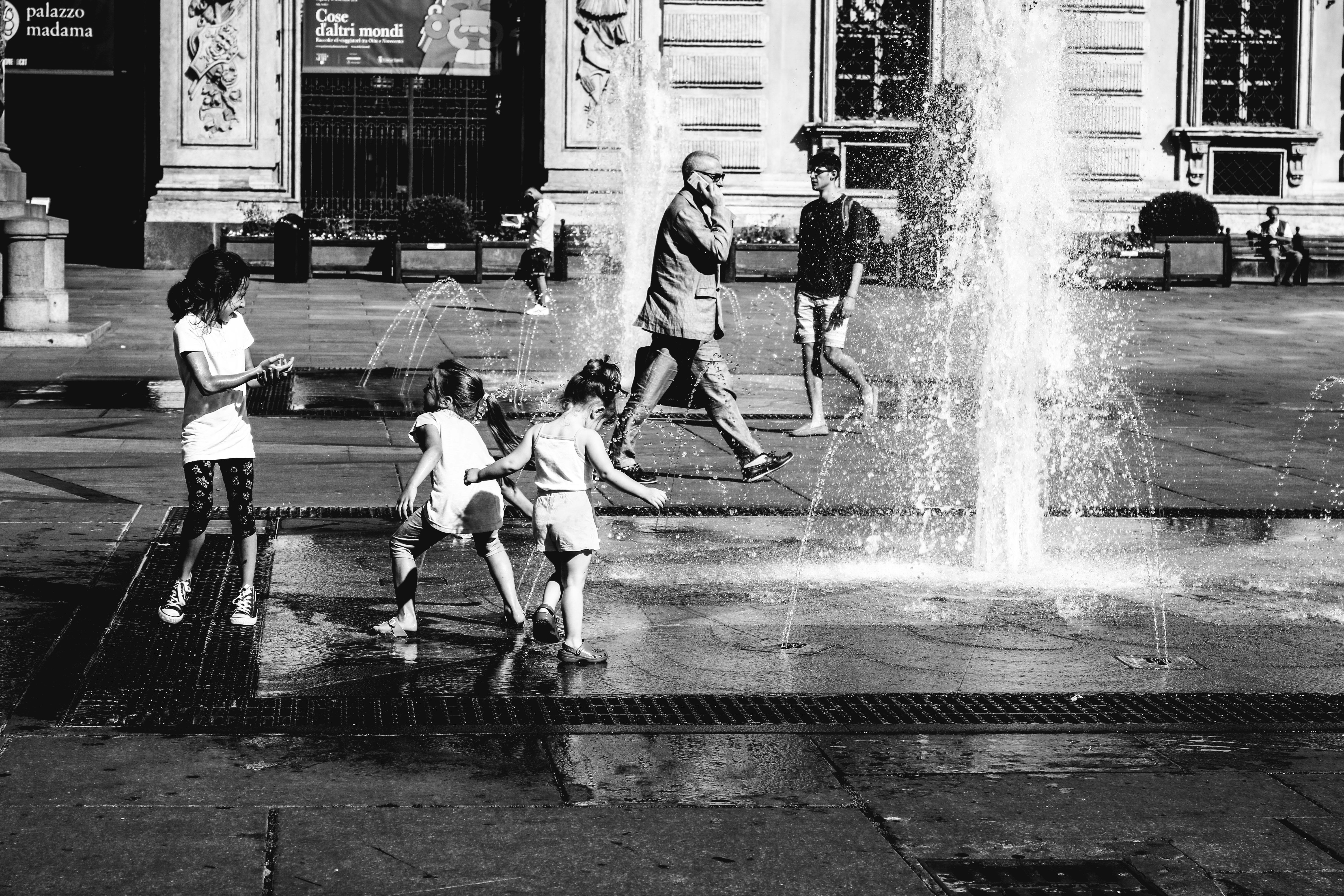 Children play near a fountain in this urban black and white street scene.