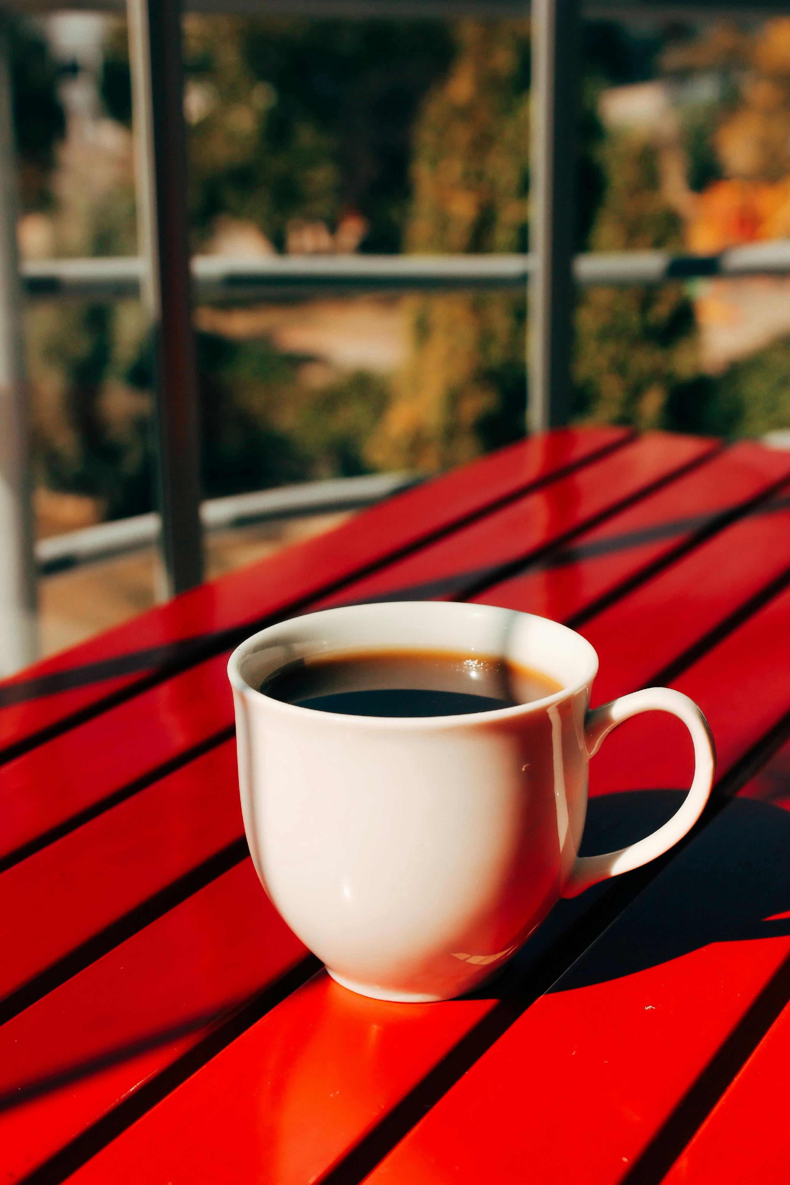 A white coffee cup on a bright red table with a blurred outdoor background, exuding a warm, inviting feel.