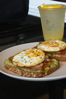 Plate of avocado toast topped with fried eggs and bacon, served with fresh lemonade.