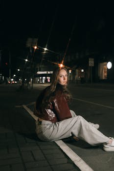 Young woman in casual outfit sitting on city street at night, illuminated by streetlights.