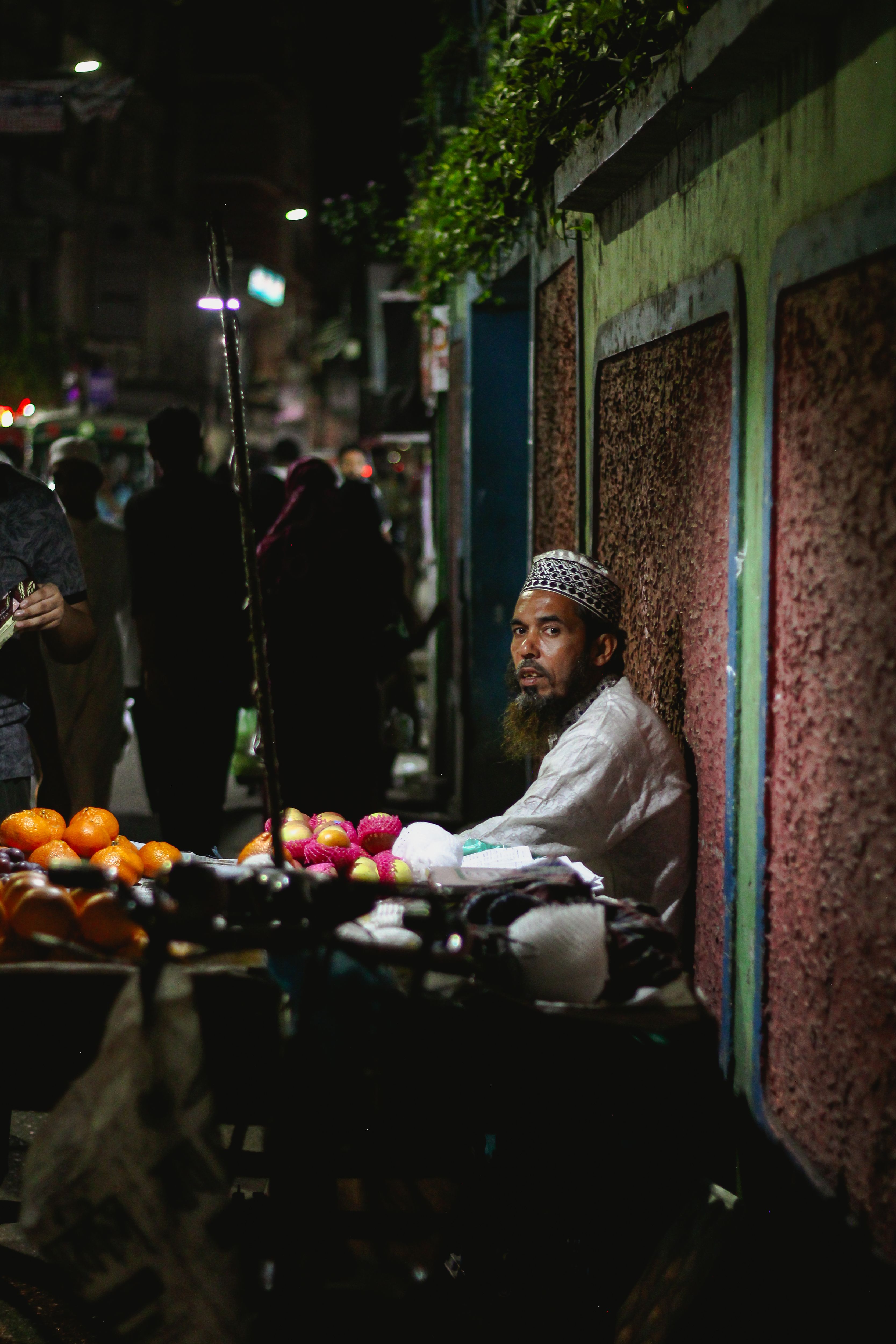 A bustling night market in Dhaka with a vendor selling fresh fruits under bright streetlights.