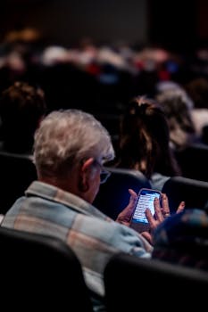 Elderly man using smartphone in dark theater, surrounded by audience.